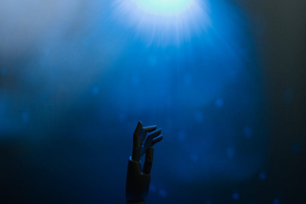 Silhouette of a robotic hand reaching towards glowing blue light in a futuristic setting.